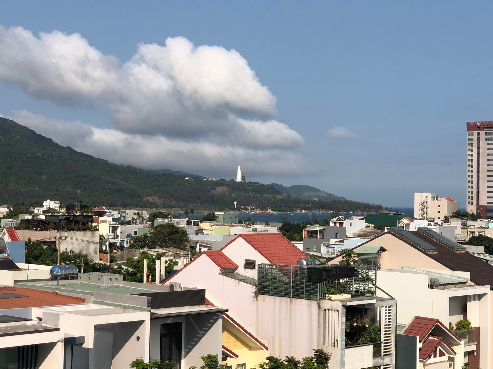 Red and white rooftops fall away toward a green ridge and the sea, with a white statue small on the far hillside under a long bank of cloud. Da Nang, the penthouse and its rooftop patio, March 2020.