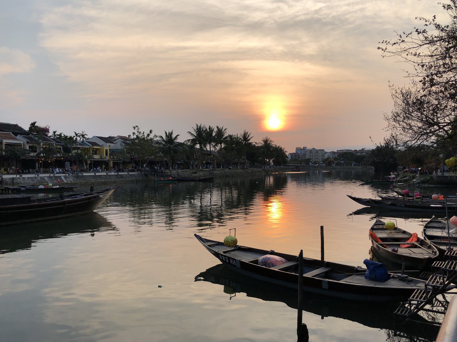 The sun sets over a still river lined with moored wooden boats, one carrying a round lantern on its bow, palms and low buildings along the far bank. Hoi An, the Thu Bon River at nightfall and a wall of silk lanterns, March 2020.
